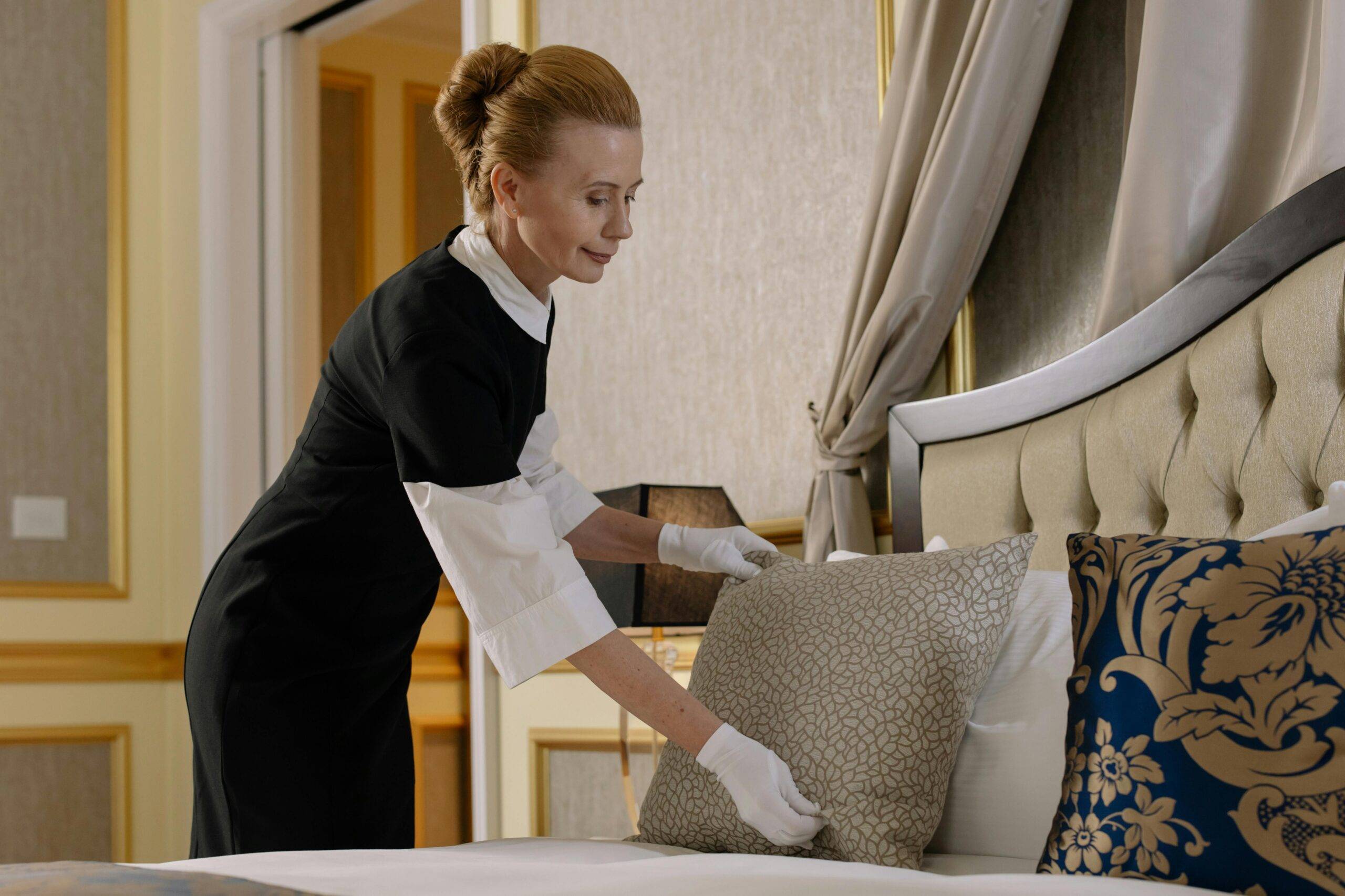 A housekeeper in uniform adjusts pillows in a luxurious hotel room, ensuring comfort and cleanliness.